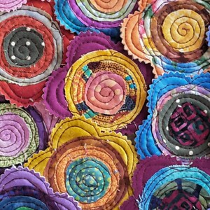 K&ouml;nnte beinhalten: Eine Sammlung von bunten Stoffkreisen mit einem Spiralmuster. Jeder Kreis besteht aus mehreren Stoffschichten, die ein strukturiertes und dreidimensionales Aussehen erzeugen. Die Kreise sind in einem zuf&auml;lligen Muster angeordnet, wodurch ein optisch ansprechendes und einzigartiges Design entsteht.