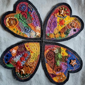 May include: Four heart-shaped fabric patches featuring a patchwork design of colourful textile scraps, intricate embroidery, and small wooden star-shaped buttons. Each heart is outlined with dark denim fabric and finished with visible stitching.