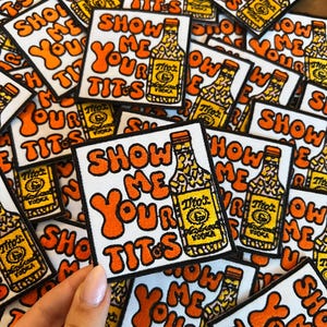 May include: A collection of white square patches with orange text that reads "SHOW ME YOUR TITS". Each patch features a yellow and black illustration of a vodka bottle. The patches are scattered.