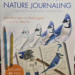 May include: A book titled "How to Teach Nature Journaling" with illustrations of blue jays and a sketch of a bird in a notebook. The cover also features two pencils and the text "Includes 31 Field Activities."