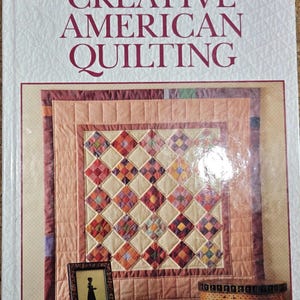 May include: A book titled "Creative American Quilting" by Better Homes and Gardens. The cover features a detailed quilt with a diamond pattern in various colors. A framed silhouette and a woven basket are also visible.