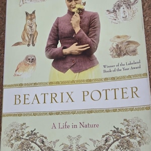 May include: A book titled Beatrix Potter: A Life in Nature by Linda Lear. The cover features a photograph of a person in a purple jacket and cap holding yellow flowers, surrounded by illustrations of a rabbit, fox, owl, mouse, sheep, and mushrooms.