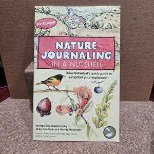 May include: A book titled "Nature Journaling in a Nutshell" with illustrations of a bird, acorn, and flowers. The cover has the text "For All Ages!" and "Draw Botanical's quick guide to jumpstart your exploration."