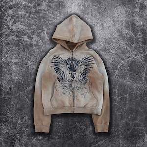 Peut inclure: Un sweat à capuche beige avec un imprimé délavé d'une créature ailée avec un crâne au centre. Le sweat-shirt a une fermeture éclair.