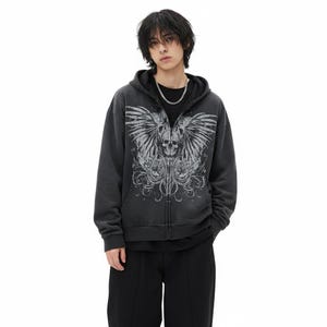 Peut inclure: Un sweat à capuche zippé gris foncé avec un motif de crâne et d'ailes sur le devant. Le motif est de couleur gris clair. Le sweat à capuche a une capuche à cordon et une fermeture éclair sur le devant. La personne porte un t-shirt noir et un pantalon noir.