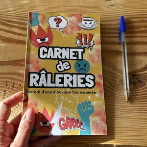 Peut inclure: Un carnet intitulé "Carnet de Râleries" avec une couverture jaune ornée de dessins animés de visages en colère, d'une tasse de café et d'un poing. Un stylo bleu est posé sur une surface en bois.