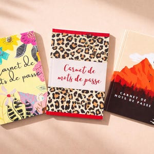 Puede incluir: Tres cuadernos con el texto "Carnet de mots de passe". Uno tiene un diseño floral, otro un estampado de leopardo y el tercero presenta un paisaje de montaña. Los cuadernos están sobre una superficie rosa claro.