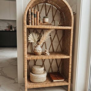 Puede incluir: Una estantería alta de mimbre color marrón claro con forma arqueada y tres niveles. El estante superior contiene cuatro libros antiguos, dos pequeñas suculentas en macetas y una lupa. El estante central presenta un jarrón de cerámica con follaje seco. El estante inferior incluye dos cajas redondas de mimbre apiladas con tapa y un diario de cuero marrón.