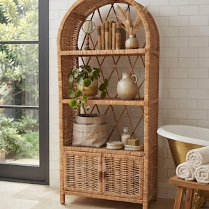 Puede incluir: Una estantería alta de mimbre color marrón claro con forma arqueada, que cuenta con tres estantes abiertos y un armario inferior con dos puertas. Los estantes contienen varios objetos decorativos, incluyendo libros, un espejo, un jarrón y una planta.