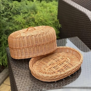 May include: An oval-shaped light brown woven wicker basket with a matching removable lid. The basket features a tight, uniform weave pattern and a small, arched handle centered on the top of the lid. The base is a separate, shallow oval tray.