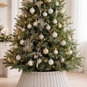 May include: A tall, green artificial Christmas tree decorated with numerous round, metallic silver and gold ornaments, white snowflake accents, and warm white string lights, standing inside a wide, tapered, light grey woven wicker tree collar.