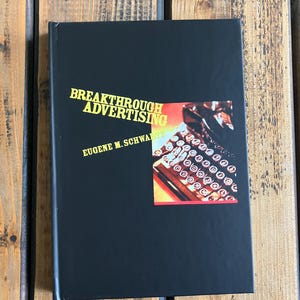 Könnte beinhalten: Ein schwarzes Buch mit dem Titel "Breakthrough Advertising" von Eugene M. Schwartz. Das Cover zeigt goldfarbene Schrift und ein farbenfrohes Bild einer Schreibmaschine. Das Buch wird auf einer Holzoberfläche präsentiert.