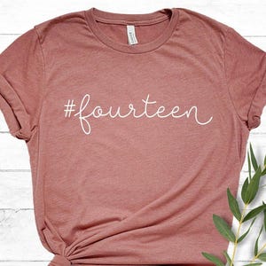 May include: A dusty rose-colored t-shirt with the word "fourteen" in white script. The shirt is knotted at the bottom and has short sleeves. A sprig of green leaves is placed on the right side of the shirt.