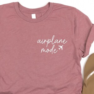 May include: A mauve t-shirt with the text 'airplane mode' in white lettering and two small airplane icons. The t-shirt is paired with denim shorts and a pair of brown flip flops.