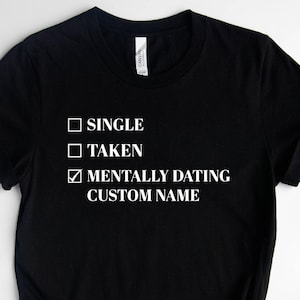 May include: Black t-shirt with a white graphic that says "Single, Taken, Mentally Dating [Custom Name]" with checkboxes next to each option. The checkbox next to "Mentally Dating" is checked.