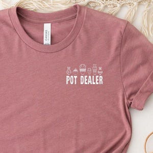 May include: A dusty rose t-shirt with the text "POT DEALER" in white, along with small plant illustrations. The shirt is knotted at the bottom and has rolled-up sleeves. The background includes decorative elements.