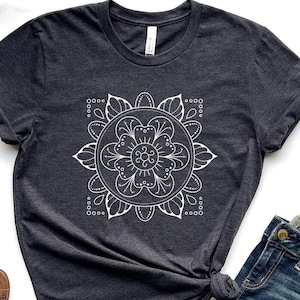 May include: Dark gray t-shirt with a white mandala design, paired with blue jeans and brown open-toe booties. The t-shirt features a round, floral-inspired pattern within a square frame. The jeans are folded neatly next to the shirt.