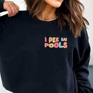 May include: Black sweatshirt with the text "I PEE IN POOLS" in colorful bubble letters. The sweatshirt is a solid black color and has long sleeves. The text is in a playful font and is centered on the chest.