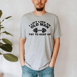 Peut inclure: Un t-shirt gris clair avec le texte "I LIFT LIKE AN OLD MAN TRY TO KEEP UP" en noir, ainsi qu'un graphique d'halt&egrave;re. Le t-shirt est port&eacute; par une personne en jean bleu.