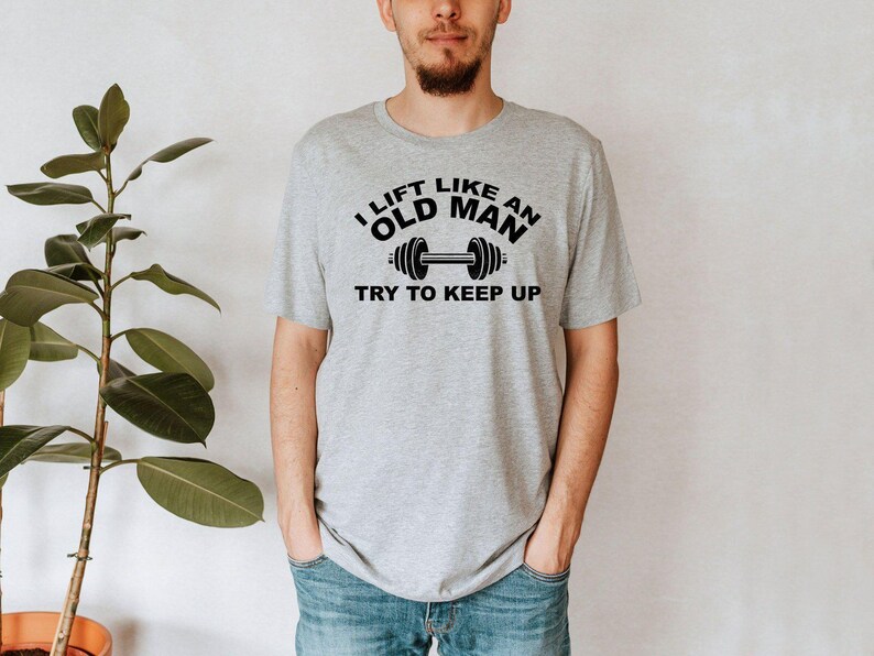 Peut inclure: Un t-shirt gris clair avec le texte "I LIFT LIKE AN OLD MAN TRY TO KEEP UP" en noir, ainsi qu'un graphique d'halt&egrave;re. Le t-shirt est port&eacute; par une personne en jean bleu.