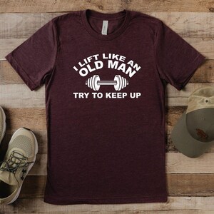 Peut inclure: Un t-shirt bordeaux avec le texte "I lift like an old man, try to keep up" en blanc. Le t-shirt est &agrave; c&ocirc;t&eacute; d'une paire de baskets beiges et d'une casquette de baseball verte et beige. Les articles sont sur une surface en bois.