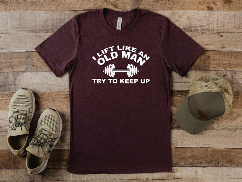 Peut inclure: Un t-shirt bordeaux avec le texte "I lift like an old man, try to keep up" en blanc. Le t-shirt est &agrave; c&ocirc;t&eacute; d'une paire de baskets beiges et d'une casquette de baseball verte et beige. Les articles sont sur une surface en bois.