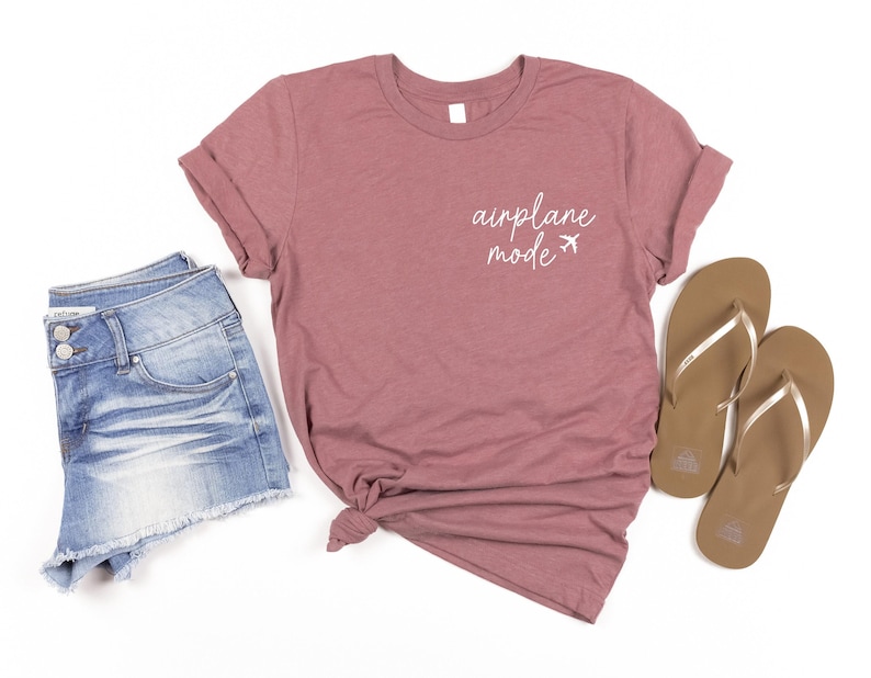 May include: A mauve t-shirt with the text 'airplane mode' in white lettering and two small airplane icons. The t-shirt is paired with denim shorts and a pair of brown flip flops.
