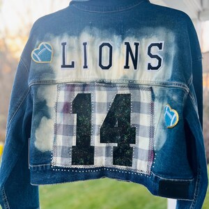 May include: A blue denim jacket with the word "LIONS" across the back. The number "14" is stitched on the back with a plaid pattern. Two heart-shaped patches with footballs are on the jacket.