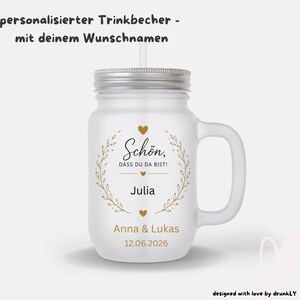 Könnte beinhalten: Ein Trinkbecher aus satiniertem Glas mit silbernem Deckel und Strohhalm. Die Vorderseite zeigt einen goldenen Lorbeerkranz, Herzen und den Text: Schön, DASS DU DA BIST!, Julia, Anna & Lukas, sowie 12.06.2026. Oben steht: personalisierter Trinkbecher - mit deinem Wunschnamen.