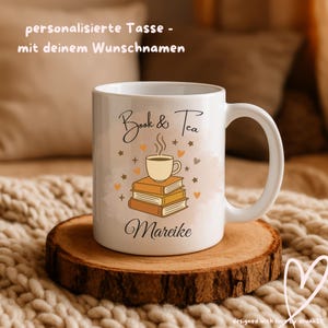 Könnte beinhalten: Weiße Keramik-Tasse mit der Aufschrift "Book & Tea" und dem Namen "Mareike". Die Tasse zeigt einen Stapel Bücher mit einer Teetasse, sowie dekorative Herzen und Sterne. Die Tasse steht auf einem Holzuntersetzer.