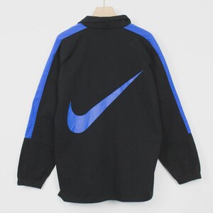 Op de afbeelding: Zwarte jas met een blauw Nike-logo en blauwe schouderstrepen. De jas heeft een kraag en lange mouwen. De Nike swoosh is groot en prominent op de achterkant.