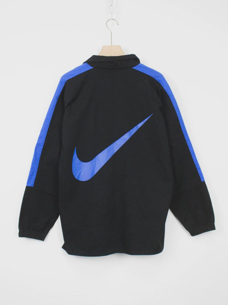 Op de afbeelding: Zwarte jas met een blauw Nike-logo en blauwe schouderstrepen. De jas heeft een kraag en lange mouwen. De Nike swoosh is groot en prominent op de achterkant.