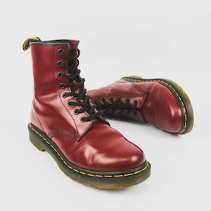 Puede incluir: Un par de botas de cuero burdeos con cordones negros. Las botas tienen un diseño clásico con detalles de costura amarilla alrededor de la suela. Las botas son un estilo de calzado popular.