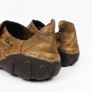 K&ouml;nnte beinhalten: Ein Paar hellbraune Lederschuhe mit Used-Optik und dunkelbraunen Gummisohlen. Die Schuhe weisen sichtbare N&auml;hte an den Seiten und an der Ferse auf, wobei der Markenname Clarks seitlich auf dem Oberleder in der N&auml;he der Fersenkappe eingepr&auml;gt ist.