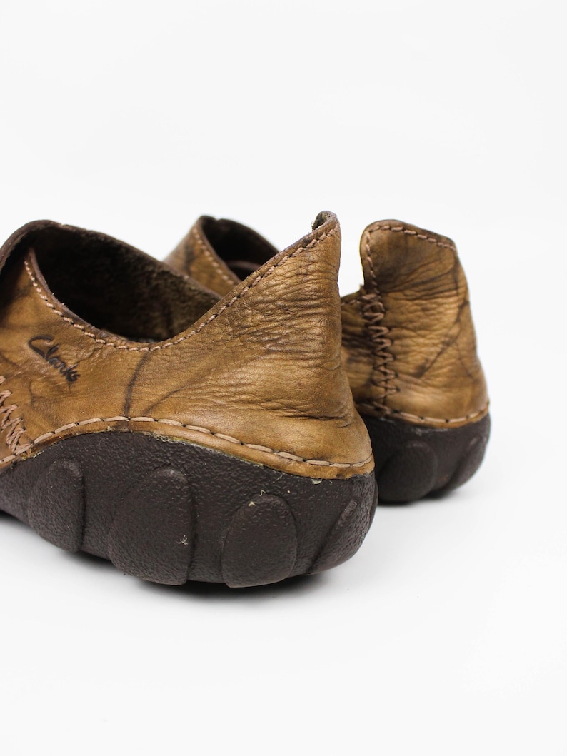 K&ouml;nnte beinhalten: Ein Paar hellbraune Lederschuhe mit Used-Optik und dunkelbraunen Gummisohlen. Die Schuhe weisen sichtbare N&auml;hte an den Seiten und an der Ferse auf, wobei der Markenname Clarks seitlich auf dem Oberleder in der N&auml;he der Fersenkappe eingepr&auml;gt ist.