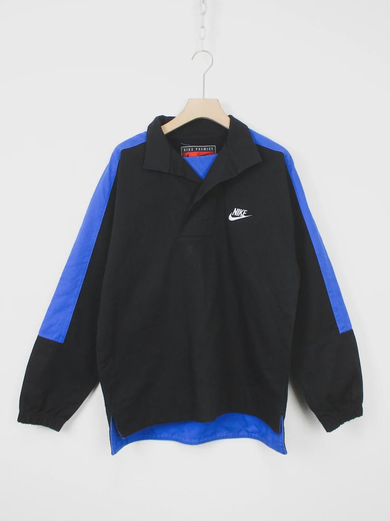 Op de afbeelding: Zwarte en blauwe Nike pulloverjas. De jas heeft een zwarte body met blauwe accenten op de mouwen en kraag. Het Nike-logo is wit op de borst geborduurd. De jas heeft een kraag en een halve ritssluiting.