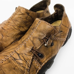 K&ouml;nnte beinhalten: Ein Paar beigefarbene Leder-Slipper mit strukturierter, marmorierter Oberfl&auml;che, markanten Zickzackn&auml;hten entlang der Kanten und einer dunkelbraunen Gummisohle. Die Schuhe verf&uuml;gen &uuml;ber ein dekoratives Schn&uuml;rdetail mit kleinen schwarzen Knebeln.