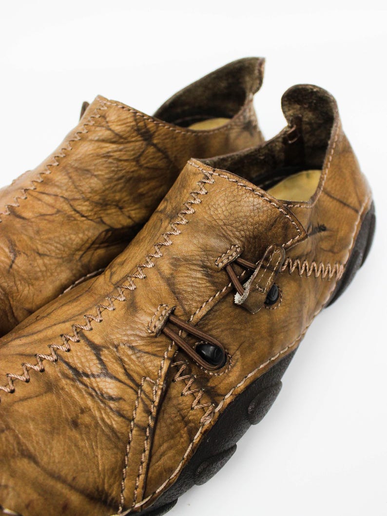 K&ouml;nnte beinhalten: Ein Paar beigefarbene Leder-Slipper mit strukturierter, marmorierter Oberfl&auml;che, markanten Zickzackn&auml;hten entlang der Kanten und einer dunkelbraunen Gummisohle. Die Schuhe verf&uuml;gen &uuml;ber ein dekoratives Schn&uuml;rdetail mit kleinen schwarzen Knebeln.