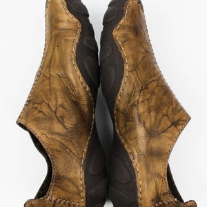 K&ouml;nnte beinhalten: Ein Paar Slipper mit einem Obermaterial aus abgenutztem, hellbraunem Leder in texturierter, marmorierter Optik. Die Schuhe haben eine dunkelbraune Gummisohle, die sich &uuml;ber Zehen und Ferse erstreckt, akzentuiert durch helle Kontrastn&auml;hte.