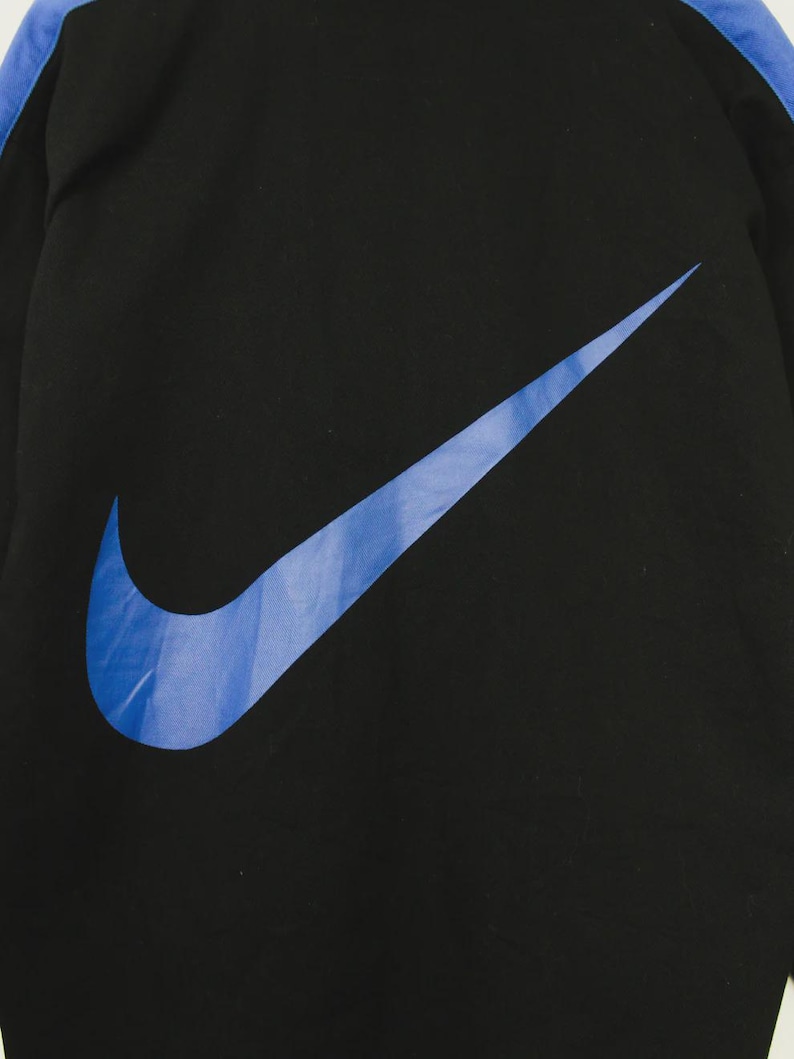 Op de afbeelding: Zwart sportjack met een groot blauw Nike swoosh-logo op de voorkant. De jas heeft blauwe accenten langs de mouwen. Het Nike-logo is effen blauw.