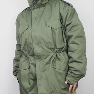 May include: Olive green military-style jacket with a high collar and multiple pockets. The jacket features a drawstring waist, a button closure, and a zipper. The jacket is made of a durable fabric.