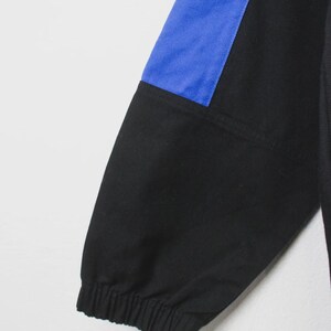 Op de afbeelding: Zwarte en blauwe mouw van een sportjack. De mouw heeft een zwarte stof met een blauw paneel op de bovenarm. De manchet is elastisch. De jas lijkt gemaakt van een duurzaam materiaal.