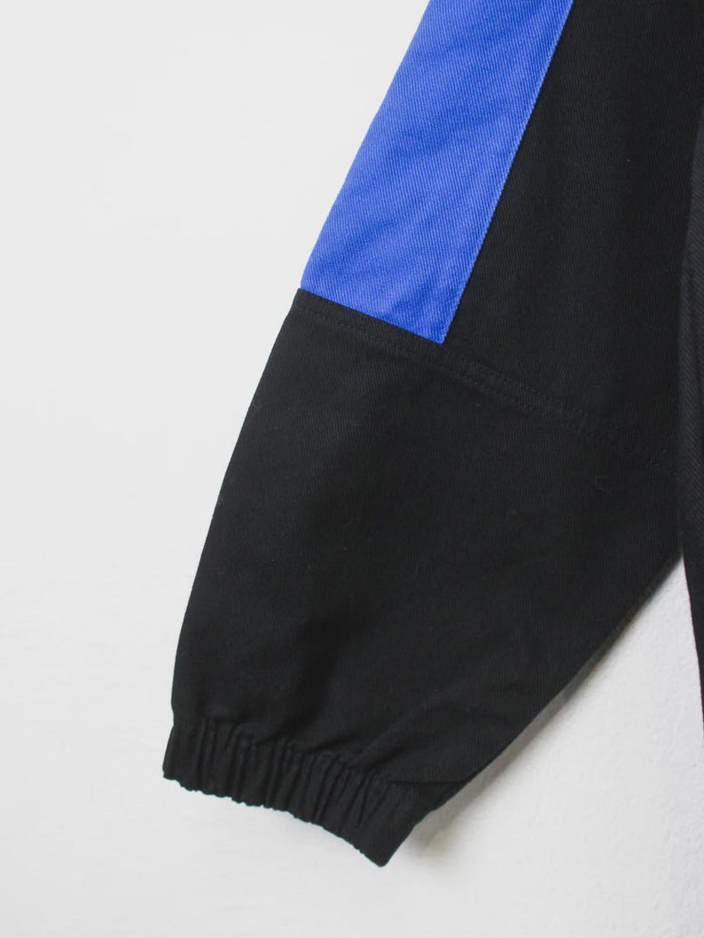 Op de afbeelding: Zwarte en blauwe mouw van een sportjack. De mouw heeft een zwarte stof met een blauw paneel op de bovenarm. De manchet is elastisch. De jas lijkt gemaakt van een duurzaam materiaal.