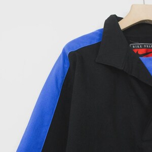 Op de afbeelding: Zwart poloshirt met een blauw schouderpaneel. De shirt heeft een kraag en een Nike Premier logo. De shirt hangt aan een houten hanger.