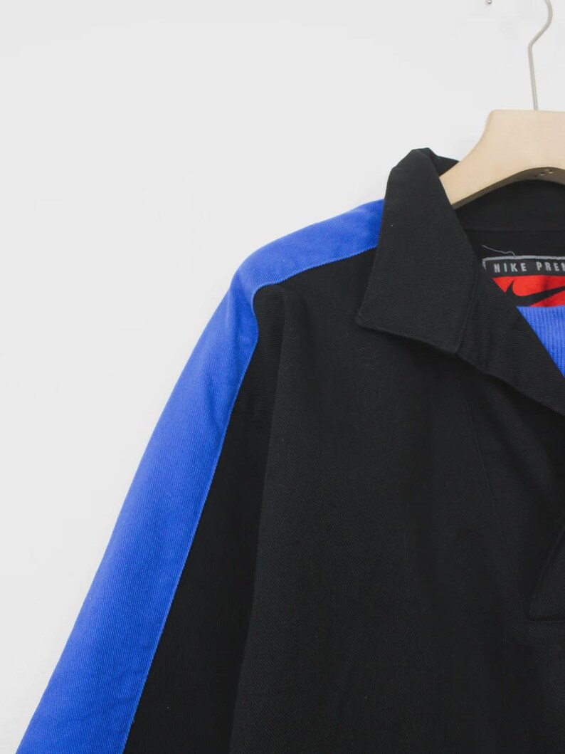 Op de afbeelding: Zwart poloshirt met een blauw schouderpaneel. De shirt heeft een kraag en een Nike Premier logo. De shirt hangt aan een houten hanger.