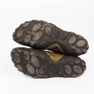 K&ouml;nnte beinhalten: Ein Paar braune Lederschuhe mit dunkelbraunen Gummisohlen, die ein ausgepr&auml;gtes Muster aus erhabenen ovalen Noppen aufweisen. Die Sohlen tragen den eingepr&auml;gten Markennamen Clarks in der Mitte und sind vor einem wei&szlig;en Hintergrund gestapelt.