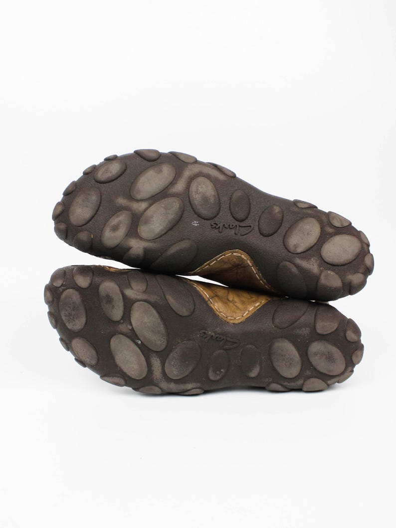 K&ouml;nnte beinhalten: Ein Paar braune Lederschuhe mit dunkelbraunen Gummisohlen, die ein ausgepr&auml;gtes Muster aus erhabenen ovalen Noppen aufweisen. Die Sohlen tragen den eingepr&auml;gten Markennamen Clarks in der Mitte und sind vor einem wei&szlig;en Hintergrund gestapelt.