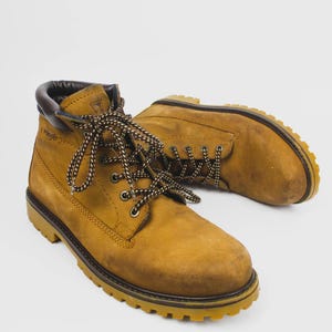 Op de afbeelding: Een paar bruin-gele werklaarzen met bruin-witte veters. De laarzen hebben een robuuste zool en een gewatteerde kraag. De merknaam "Wrangler" is zichtbaar aan de zijkant. Gemaakt van duurzaam materiaal.