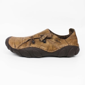 K&ouml;nnte beinhalten: Ein beigefarbener Schlupfschuh aus Leder mit Used-Optik, ausgestattet mit einer dunkelbraunen, strukturierten Gummisohle, einer abgerundeten Schuhspitze und einem dekorativen Schn&uuml;rdetail auf der Oberseite. Der Markenname Clarks ist seitlich eingepr&auml;gt.