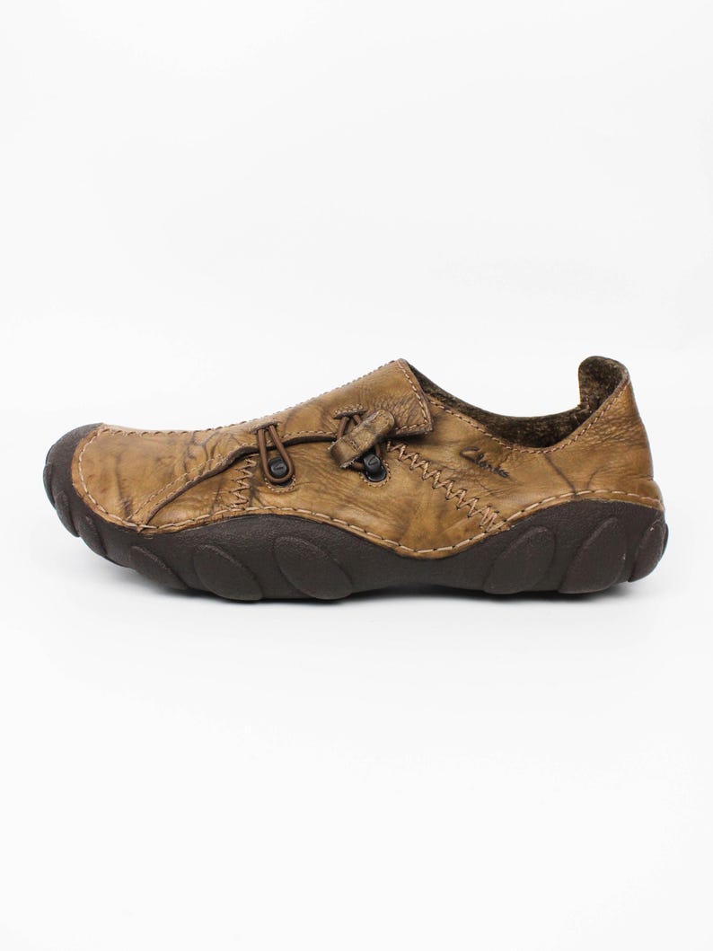 K&ouml;nnte beinhalten: Ein beigefarbener Schlupfschuh aus Leder mit Used-Optik, ausgestattet mit einer dunkelbraunen, strukturierten Gummisohle, einer abgerundeten Schuhspitze und einem dekorativen Schn&uuml;rdetail auf der Oberseite. Der Markenname Clarks ist seitlich eingepr&auml;gt.